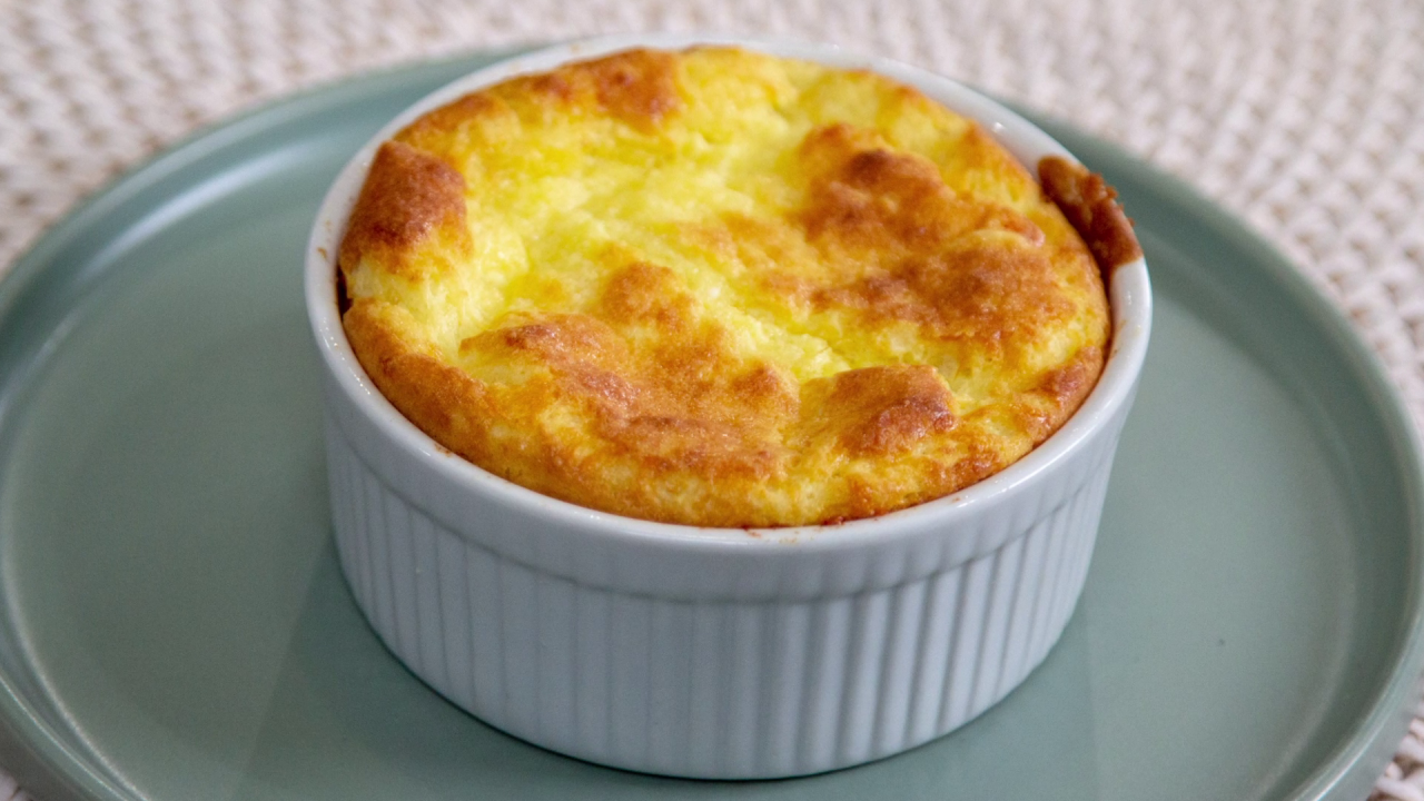 One of the finished Mini Cheese Souffles, served in the ramekin it was baked in atop a greyish-blue plate.