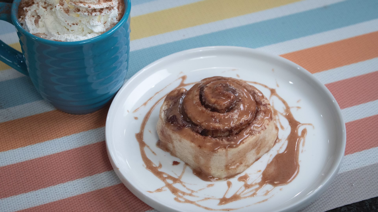 An Apple Pie Cinnamon roll, covered in frosting, sits on a white plate on a decorative place mat. A mug of the Pumpkin Spice Hot Cocoa, topped with whipped cream, is next to the plate, barely in frame in the top left corner.