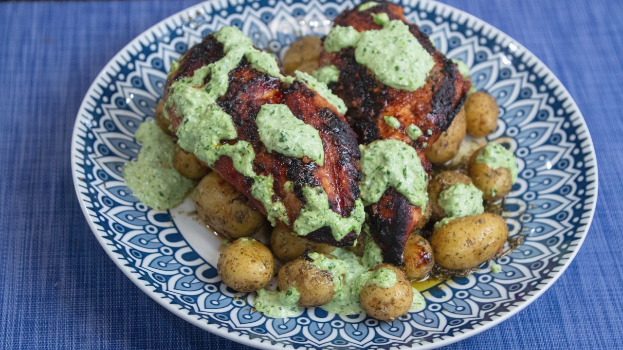 The finished Peruvian Chicken, served on top of roasted potatoes with the Green Sauce drizzled over it. It's served in a blue plate with a peacock pattern on a blue place mat.