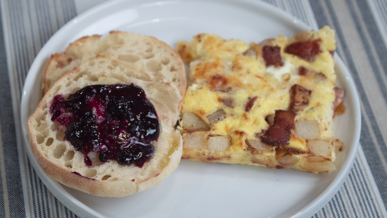 A serving of the Bacon and Potato Breakfast Bars on a white plate next to two halves of an English muffin, stacked on top each other, on the left side of the image. The top half has jelly spread on it.