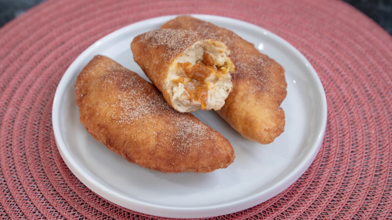 The finished Fried Pumpkin Cheesecake Hand Pies, dusted with cinnamon and sugar, sitting on a white plate. One pie is cut open to show the gooey interior.