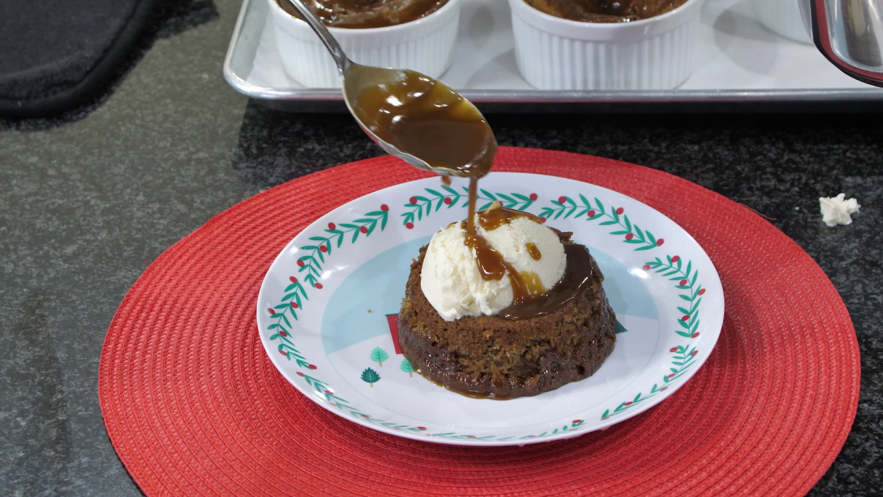 The finished pudding, served on a white plate with a holiday design, topped with vanilla ice cream. A ladle drizzles the toffee sauce onto it.