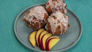 The finished peach fritters, drizzled with the glaze, served on a bluish-gray plate with freshly sliced peaches on the side. The plate sits atop a turquoise placemat.