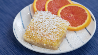 A slice of the Orange Bars, dusted with powdered sugar and served with three slices of blood oranges on a white plate with thin blue stripes atop of blue tablecloth.