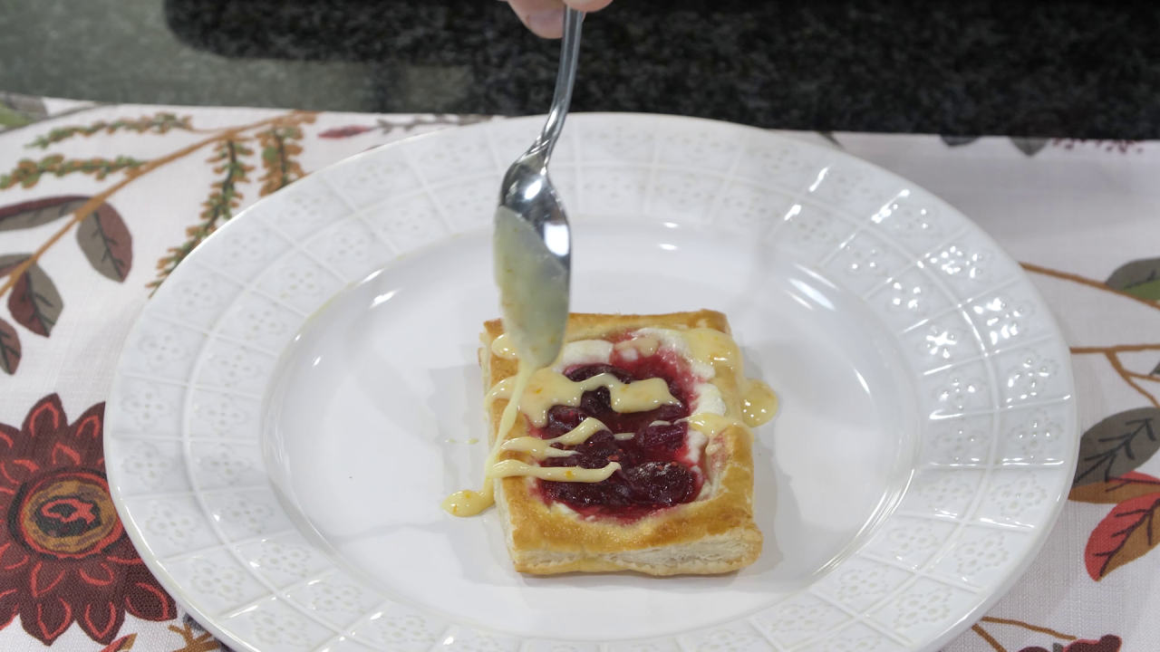 One of the freshly baked danishes, sitting on a white plate. A spoon drizzles the orange glaze over it.